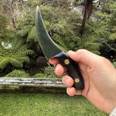 Hand holding a knife with a green blade and black handle against a natural background